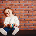 Cute little girl sitting in a room. Child have fun at home near wall. Little girl is studying a colorful cube